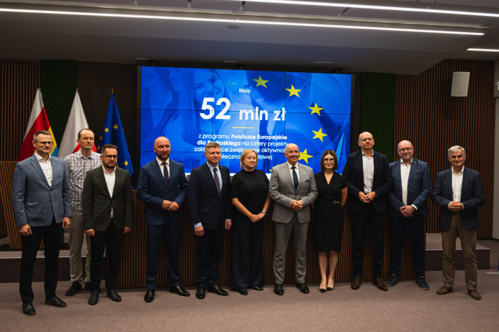 Grupa osób, kobiet i mężczyzn stoi na tle flag Polski i województwa podlaskiego oraz slajdu z kwotą dofinansowania projektów.
