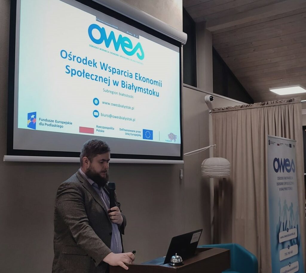 Mężczyzna z brodą mówi do mikrofonu. W tle prezentacja dotycząca OWES wyświetlona na ekranie.