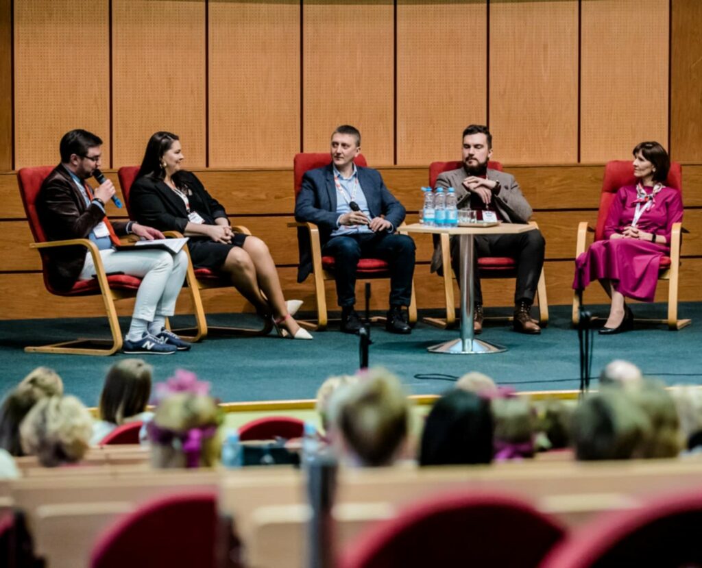 Pięć osób, kobiet i mężczyzn siedzi na krzesłach na scenie. Przygląda im się ludzie siedzący na widowni.