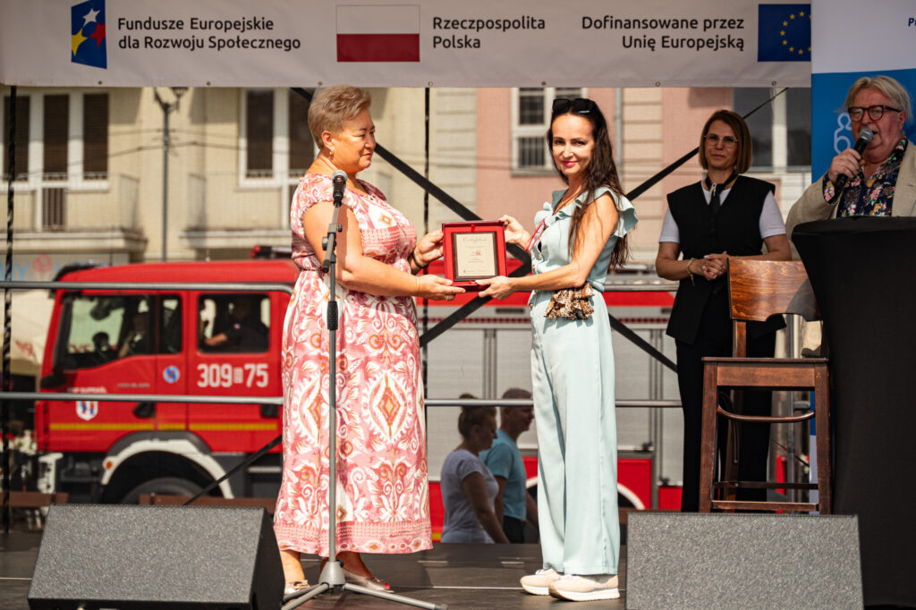 Dwie kobiety stoją na scenie. Jedna z nich wręcza certyfikat.