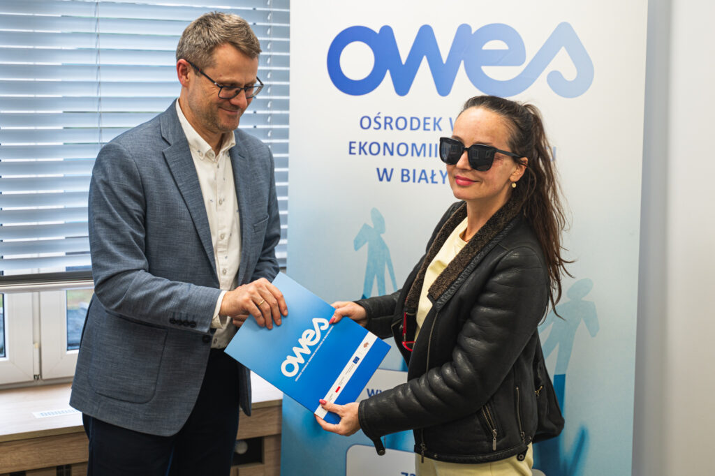 Mężczyzna podaje kobiecie w okularach teczkę z umową.