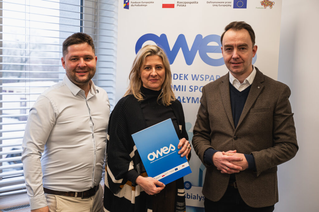 Na zdjęciu stoi dwóch mężczyzn i kobieta, która trzyma w ręku teczkę z umową.