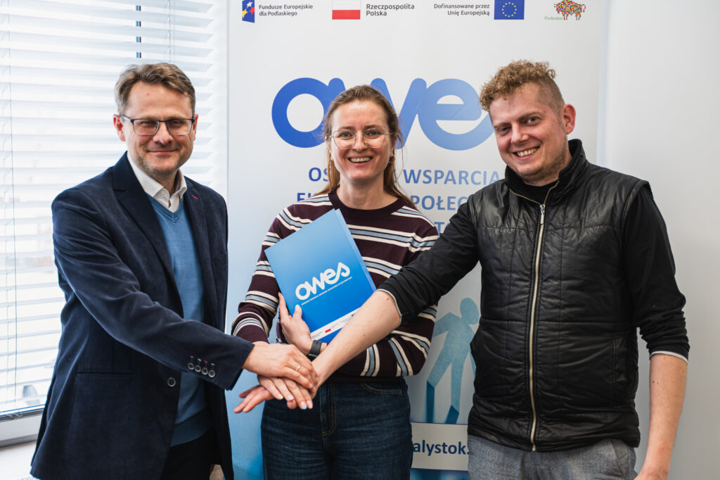 Dwaj mężczyźni i kobieta podają sobie ręce. Kobieta trzyma w ręku umowę.