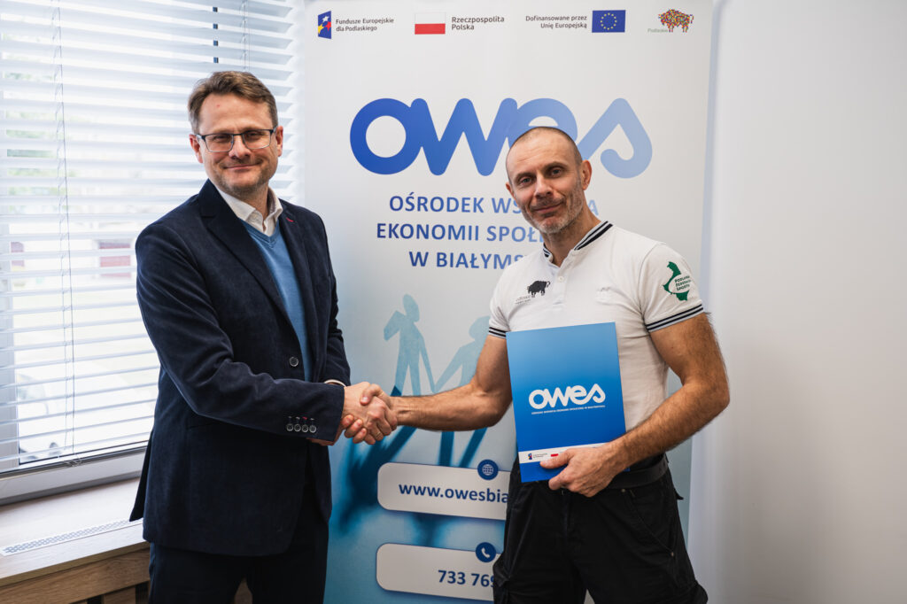 Dwaj mężczyźni podają sobie ręce. Mężczyzna po prawej trzyma w ręku umowę.