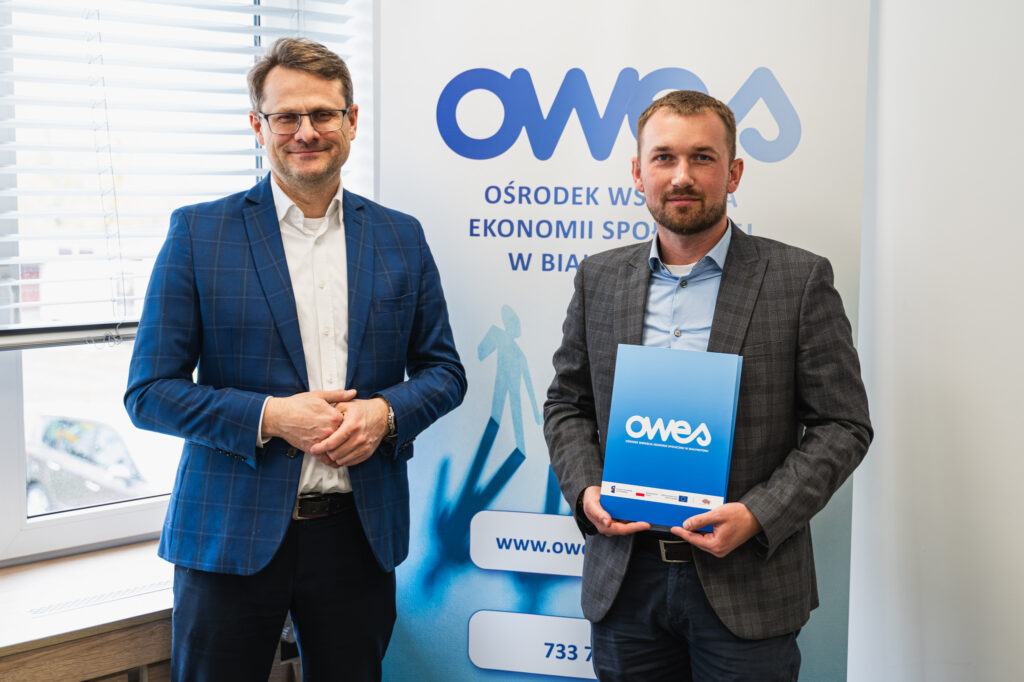 Dwóch uśmiechniętych mężczyzn stoi na tle ścianki z napisem OWES. Mężczyzna po prawej stronie trzyma teczkę z umową.