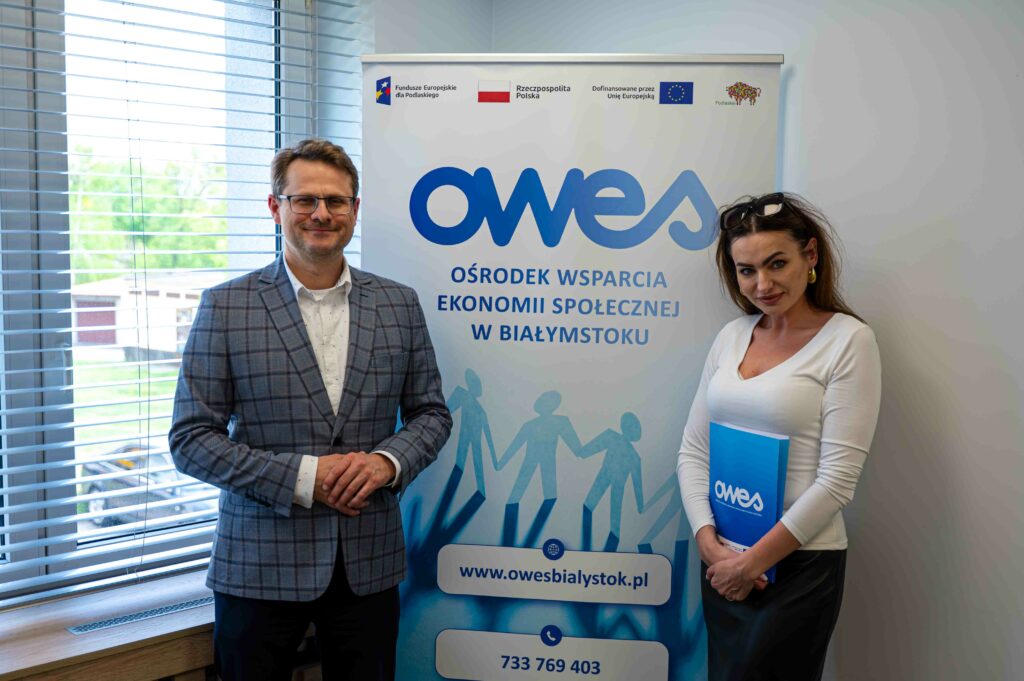 Mężczyzna i kobieta stoją na tle bannera OWES. Kobieta trzyma w rękach teczkę z umową.
