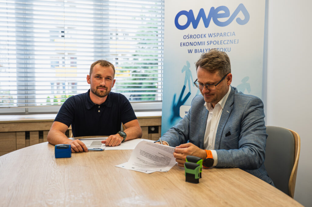 Dwóch mężczyzn siedzi przy stole, jeden z nich podpisuje aneks do umowy.