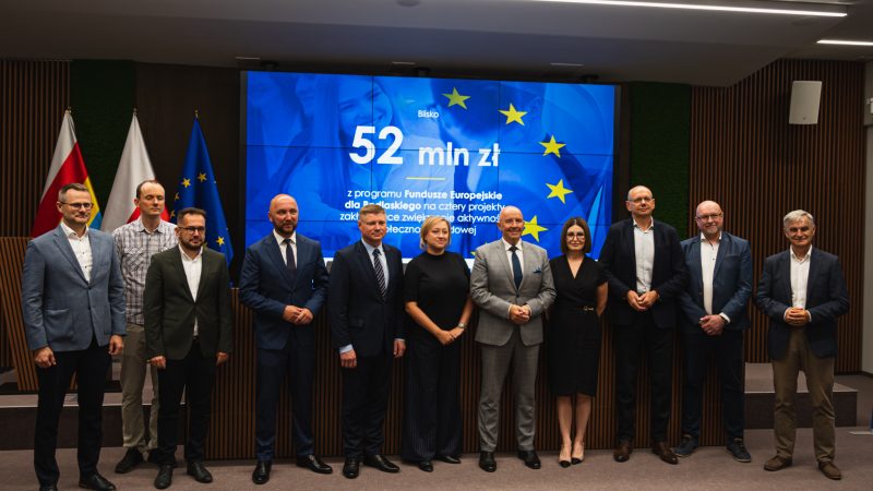 Grupa osób, kobiet i mężczyzn stoi na tle flag Polski i województwa podlaskiego oraz slajdu z kwotą dofinansowania projektów.