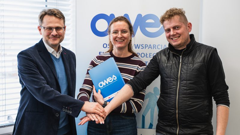 Dwaj mężczyźni i kobieta podają sobie ręce. Kobieta trzyma w ręku umowę.