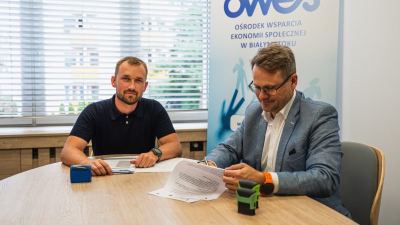 Dwóch mężczyzn siedzi przy stole, jeden z nich podpisuje aneks do umowy.