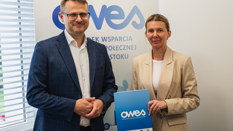 Mężczyzna i kobieta stoją na tle rollupu z napisem OWES. Kobieta trzyma w dłoni teczkę z umową.
