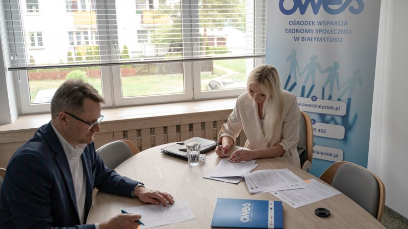 Mężczyzna i kobieta z jasnymi włosami siedzą przy stole i podpisują dokumenty. Na dalszym tle roll-up z logotypem OWESu.