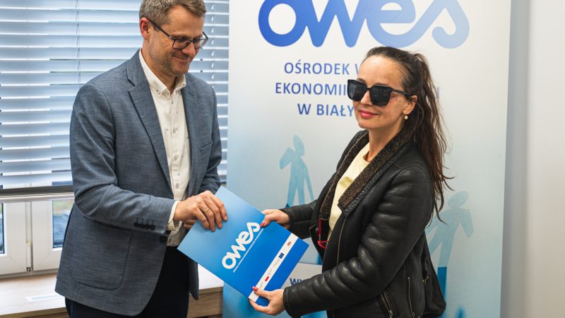 NilSine4 Mężczyzna podaje kobiecie w okularach teczkę z umową.