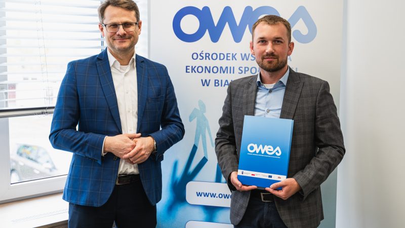 Dwóch uśmiechniętych mężczyzn stoi na tle ścianki z napisem OWES. Mężczyzna po prawej stronie trzyma teczkę z umową.