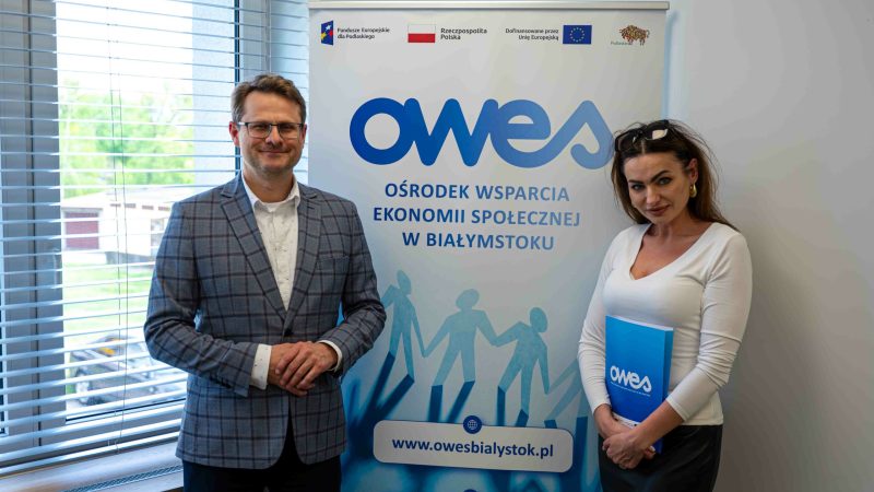 Mężczyzna i kobieta stoją na tle bannera OWES. Kobieta trzyma w rękach teczkę z umową.
