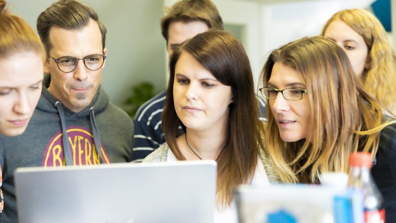 Grupa osób, kobiet i mężczyzn wpatruje się w ekran laptopa.